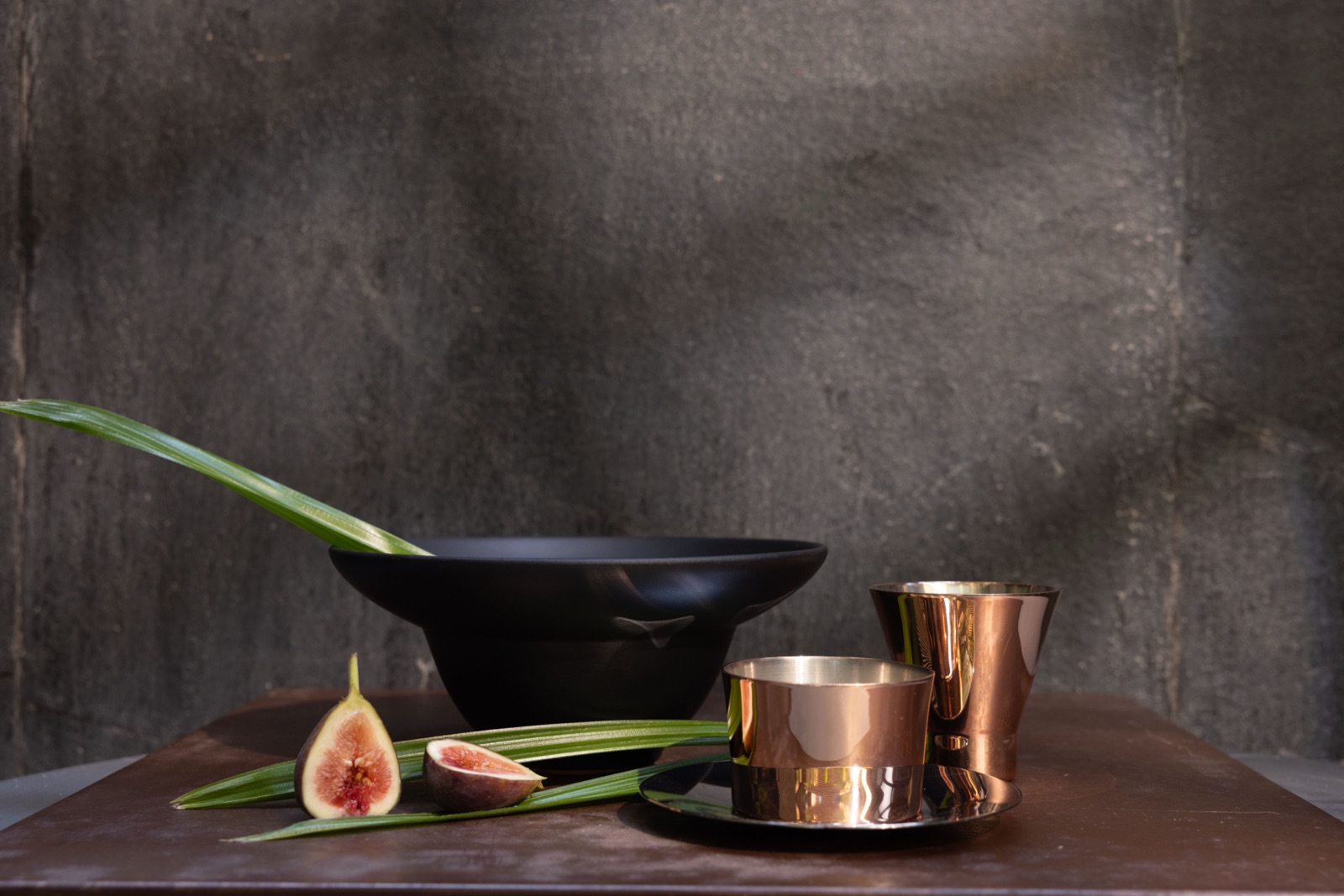 A black Iteru stoneware bowl and copper Meteorite tumblers on a dark table, with a cut fig and a palm leaf